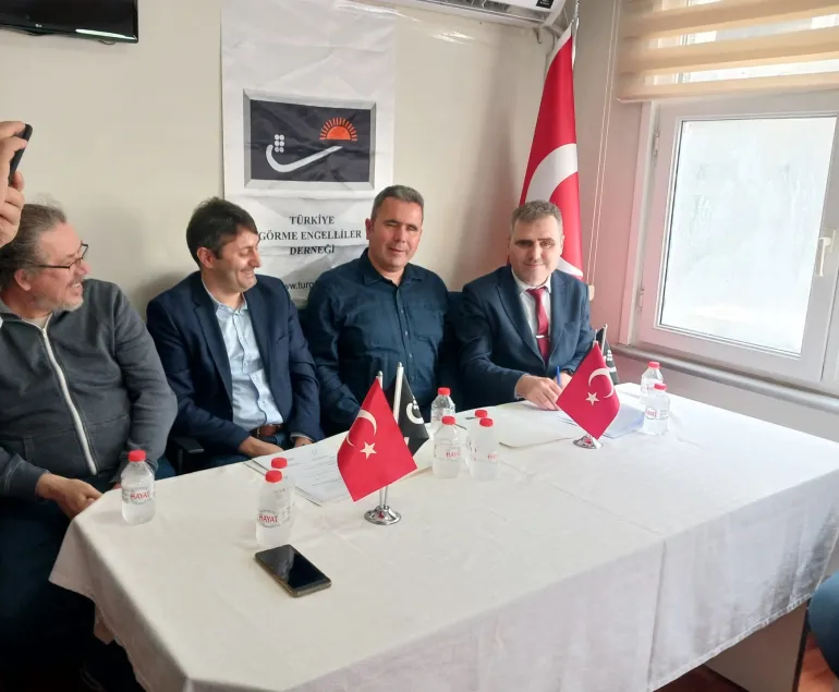 : Protokol imza töreni fotoğrafı. Beyaz masa örtülü protokol masasında soldan sağa dört erkek yetkili oturuyor. Masanın üzerinde imzalanacak belgeler, su şişeleri, siyah bir cep telefonu ve iki adet Türk bayrağı bulunuyor. Arka planda beyaz duvarda TURGED logosu ve "Türkiye Görme Engelliler Derneği" yazısı, yanında Türk bayrağı ve beyaz tahta görülüyor.