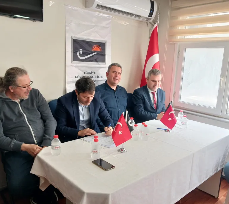 Türkiye Görme Engelliler Derneği'nde düzenlenen protokol imza töreninde, masada oturan dört erkek temsilciden biri protokolü imzalarken, diğerleri yanında oturuyor; masada Türk bayrakları, su şişeleri ve imza evrakları bulunuyor, arka planda dernek logosu ve Türk bayrağı yer alıyor.