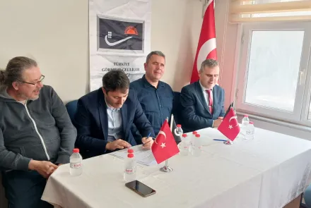 Türkiye Görme Engelliler Derneği'nde düzenlenen protokol imza töreninde, masada oturan dört erkek temsilciden biri protokolü imzalarken, diğerleri yanında oturuyor; masada Türk bayrakları, su şişeleri ve imza evrakları bulunuyor, arka planda dernek logosu ve Türk bayrağı yer alıyor.